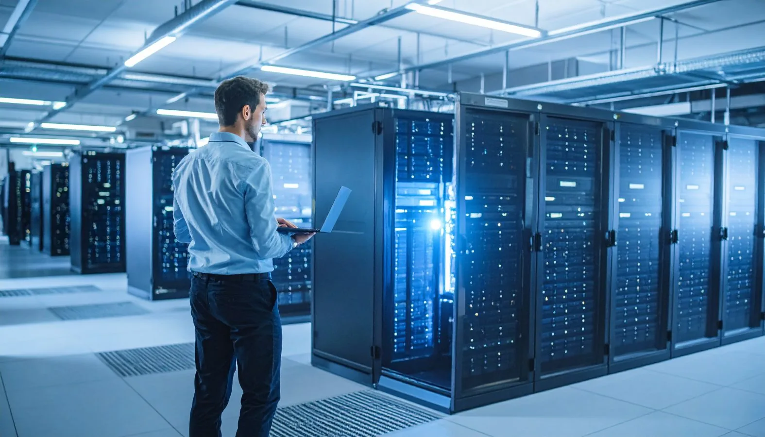 An IT professional holding a laptop while monitoring server racks in a modern data center.